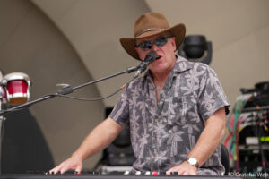 Musician wearing a brown hat and sunglasses sings into a microphone while playing a keyboard on stage.