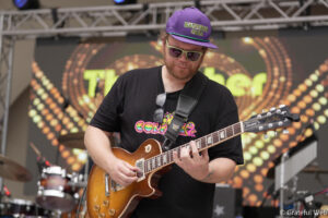 Guitarist wearing a purple hat and light-green sunglasses plays a sunburst electric guitar on a festival stage with a colorful backdrop behind him.