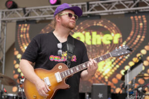 Guitarist in a purple cap and lime-green sunglasses playing a sunburst electric guitar on a stage backdrop.