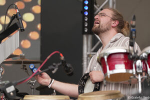 Drummer wearing a striped shirt plays a red drum kit on stage with a microphone in front of him.