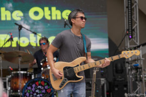 Male bass guitarist wearing sunglasses and a gray T-shirt plays a natural wood Fender bass on an outdoor stage with drums in the background.