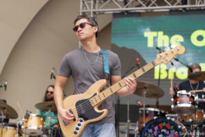 Male bassist wearing sunglasses and a gray T-shirt plays a natural wood bass on stage during a live concert.