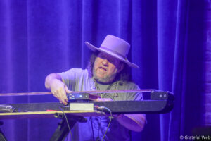 Musician wearing a light hat and gray shirt playing a pedal steel guitar on a stage with purple curtains behind him.