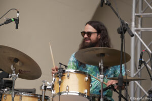 Drummer with long hair and beard wearing sunglasses plays a beige drum set amid multiple microphones on stage.