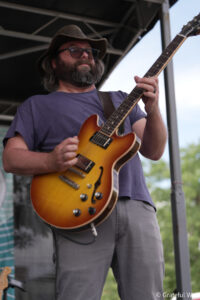 Bearded guitarist wearing a brown hat and glasses playing a sunburst semi-hollow electric guitar on an outdoor stage.] ,(