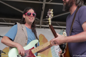 Musician wearing bright pink sunglasses plays a mint-green bass guitar on stage, another guitarist beside him.