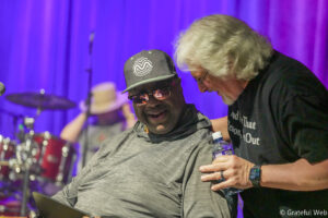 Two men on a stage sharing a smile during a performance; one wears a gray cap with a white wave logo and sunglasses, the other with white hair leans in.