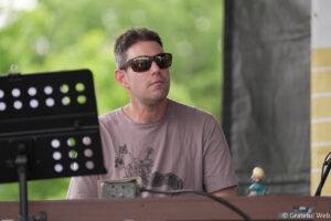 Man wearing sunglasses sits at a music setup outdoors, with a black perforated music stand in front and greenery in the blurred background.