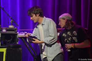 Two men on a stage with purple lighting; one in a denim shirt adjusting headphones near DJ gear, the other in a bandana watching.
