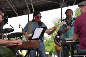 Musician with long dark hair and bright pink sunglasses plays a mint-green bass on an outdoor stage while others perform nearby.