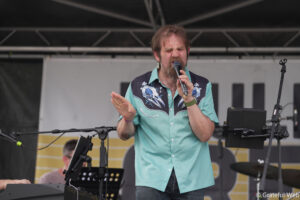 Male singer on stage, eyes closed, singing into a handheld microphone with a turquoise shirt and floral pattern backdrop behind him.