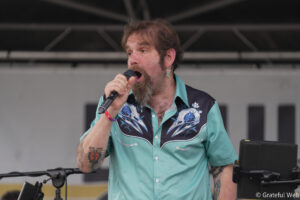 Male singer with a beard and earrings singing into a microphone on stage, wearing a turquoise floral shirt.