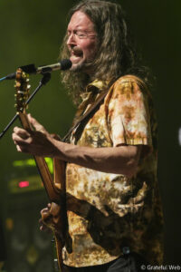 Male guitarist singing into a microphone on stage, wearing a tie-dye shirt and playing an acoustic guitar into a stand mic.