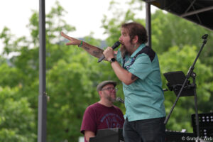 Lead singer on an outdoor stage, wearing a teal shirt and microphone in hand, with a keyboard player in the background and green trees behind them.