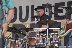 Drummer in a black cap and mirrored sunglasses plays a red drum kit on a stage with microphones and a backdrop banner behind him.