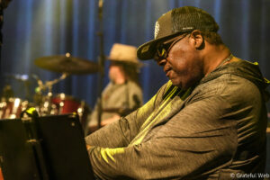 Close-up of a Black man wearing a striped hoodie, cap and sunglasses, playing keyboard on stage with drums blurred in the background.