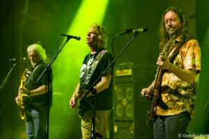 Three musicians on a smoky stage, each with an electric guitar, performing into microphones under green stage lights.
