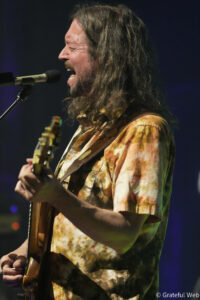 Male musician with long hair singing into a microphone while playing an electric guitar on stage, wearing a tie-dye shirt.