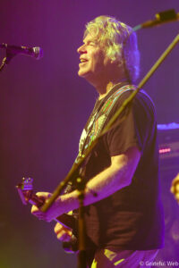 Side-profile of a guitarist singing into a microphone on stage, wearing a black shirt and a colorful guitar strap.
