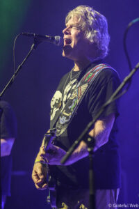 Male guitarist singing into a microphone on stage, wearing a graphic t-shirt and a colorful guitar strap under blue lighting.