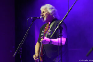 Musician with white hair and glasses singing into a microphone while playing an electric guitar on a purple-lit stage.
