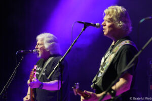 Two male musicians perform on stage under purple lighting: one with white hair singing into a microphone, the other playing a patterned guitar beside him.