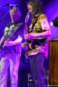 Two male guitarists perform on a purple-lit stage; one wears a black graphic tee, the other a tie-dye shirt with long hair.