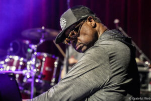 Male musician wearing a gray shirt, black cap, and sunglasses on stage with drums and purple lights in the background.
