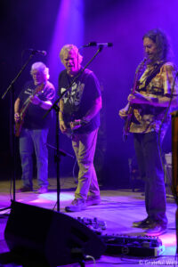 Three guitarists performing on a purple-lit stage, singer center with microphones in front and pedalboard at their feet.