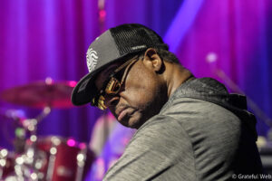 Close-up of a man in a gray shirt wearing a black trucker cap and gold sunglasses on a purple-lit stage, looking downward.