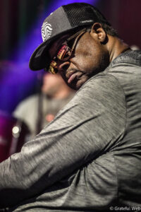 Close-up of a man wearing a black cap with a RYTHM label and mirrored sunglasses, looking down on stage amid purple lighting.