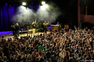 Crowd of concertgoers with hands raised toward a band performing on a smoky stage under blue and white lights.