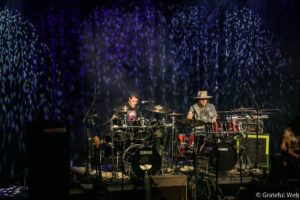 Two drummers on a dim stage playing a Yamaha drum kit under purple, patterned lights.