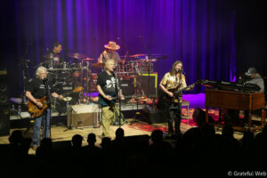 Small-stage rock band performing under purple lights; five musicians with drums, keyboards, guitars and vocals, facing the crowd.