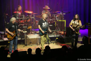 Four musicians perform on a dimly lit stage: two guitarists, a drummer, and a singer, with purple stage lights in the background.