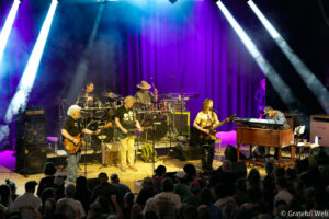 Live band on stage performing under colorful purple and blue lights, with guitarists, drummer, keyboardist, and a singer; audience watches from below.