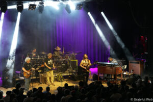 Live concert scene with musicians playing guitars, drums, and keyboards on a dimly lit stage under purple and white spotlights, audience in foreground.