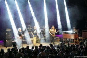 Band performs on a lit stage with four musicians; white spotlights shine downward as a crowd watches from the foreground.