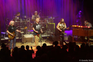 Band performs live on a lit stage: guitarist vocals center, guitarist right, drummer back, keyboardist far right, purple and red lighting, cheering audience.