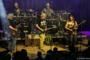 Band performing on stage with drummer in back and three guitarists upfront; purple stage lights and an attentive audience in the foreground.