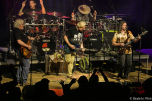 Band performs live on stage: drummer in back with Yamaha drums, and three guitarists front and center, audience hands raised.