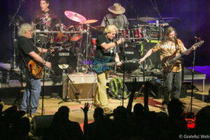 Band of four musicians on stage playing guitars and drums during a live performance, with audience silhouettes in the foreground.