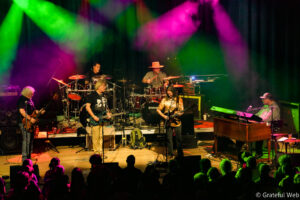 Live band performing on stage with vibrant green and magenta lights; guitarist front center, drummer behind, keyboardist to the right, audience in foreground.