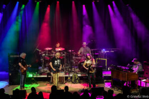 A rock band performing on a brightly lit stage with colorful red, blue, green, and purple spotlights and musicians playing guitars and drums.