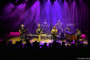 Live band on stage under purple spotlights, guitarist and singer at front with drummer and keyboard player in the back, audience silhouettes in foreground.