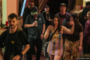 Woman in a colorful sleeveless dress raises a drink with a straw while surrounded by friends at a dimly lit party bar.