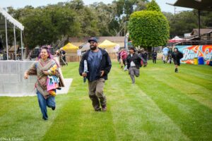 People running toward the camera at an outdoor festival in a grassy park with colorful tents in the background.