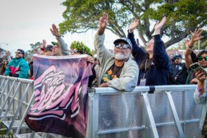 Crowd of diverse fans behind a metal barricade at an outdoor event, many raising their hands in celebration.