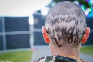 Back of a person’s head with a geometric buzz-cut pattern, outdoors at dusk with blurred lights in the background