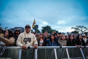 Crowd of young concertgoers pressed against a barrier at an outdoor festival under a cloudy blue sky.